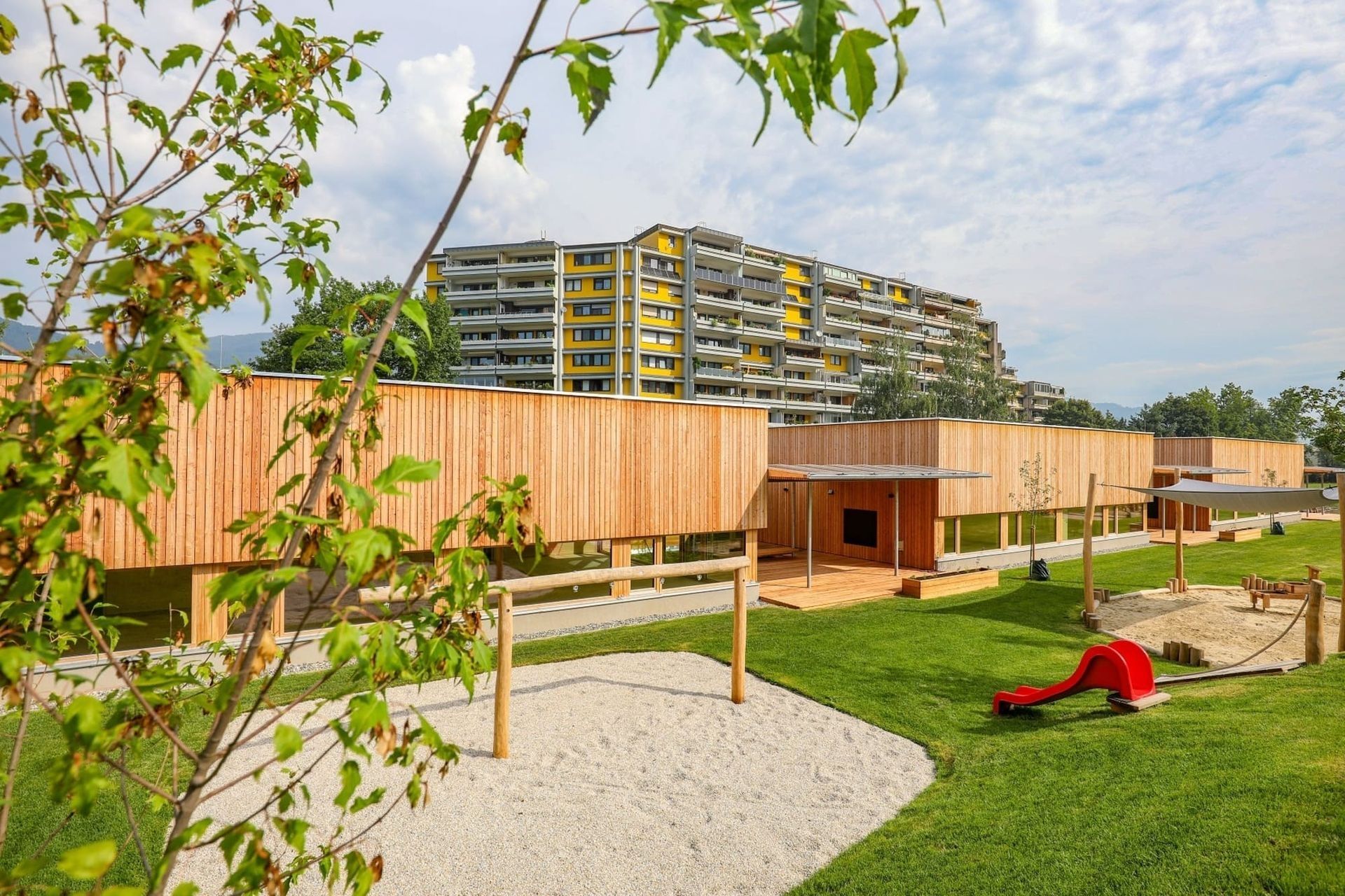 Modernes Holzgebäude mit einer überdachten Terrasse, umgeben von Gras und einem Sandkasten. In der Nähe befindet sich ein mehrstöckiges Wohnhaus mit bunten Balkonen.