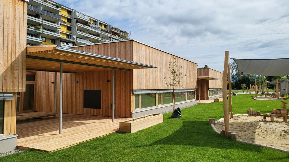 Modernes Holzgebäude mit einer überdachten Terrasse, umgeben von Gras und einem Sandkasten. In der Nähe befindet sich ein mehrstöckiges Wohnhaus mit bunten Balkonen.