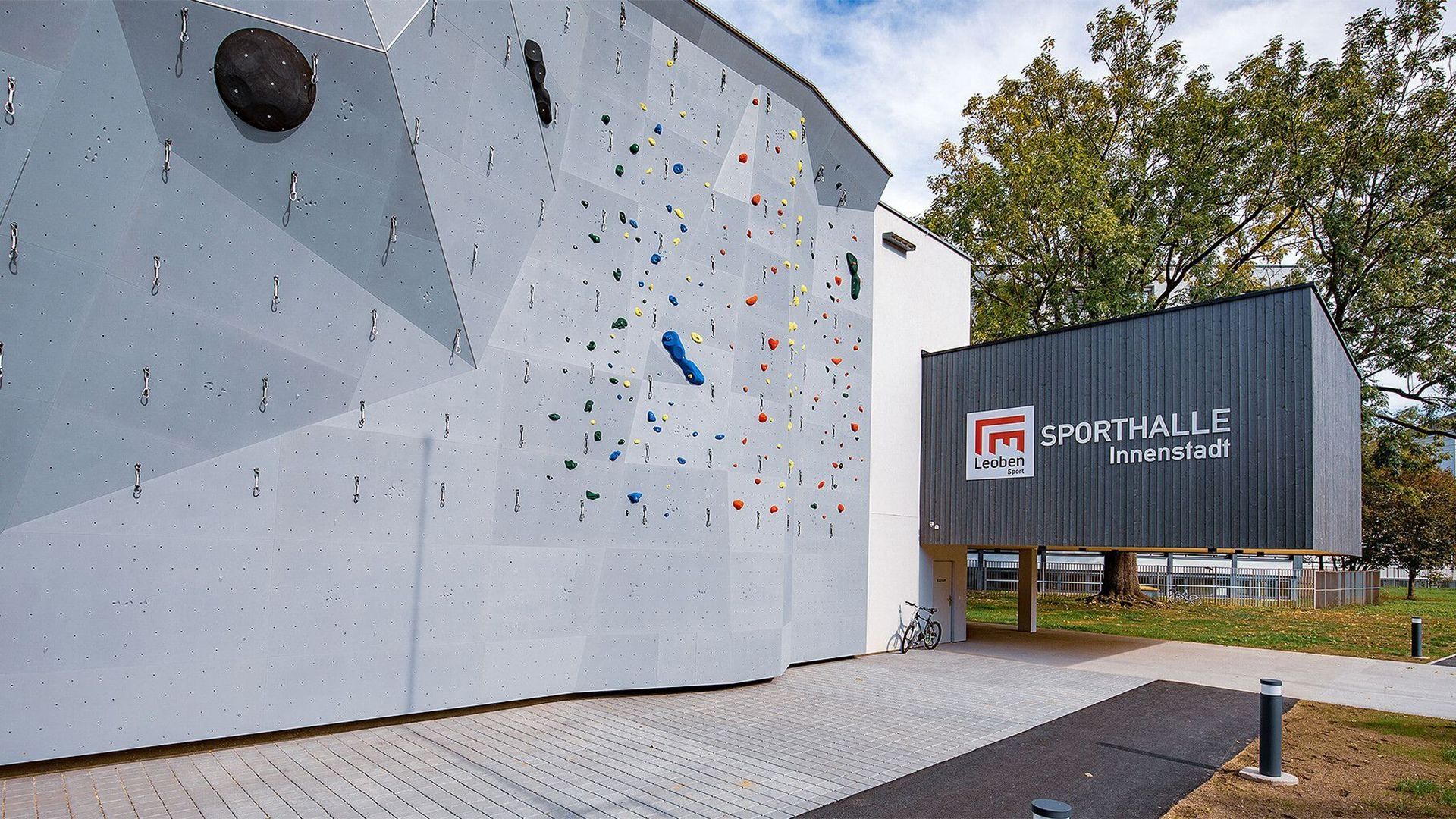 Kletterwand und Turnhalle im Freien mit einem Schild Sporthalle Innenstadt. Bäume im Hintergrund und ein gepflasterter Weg davor.