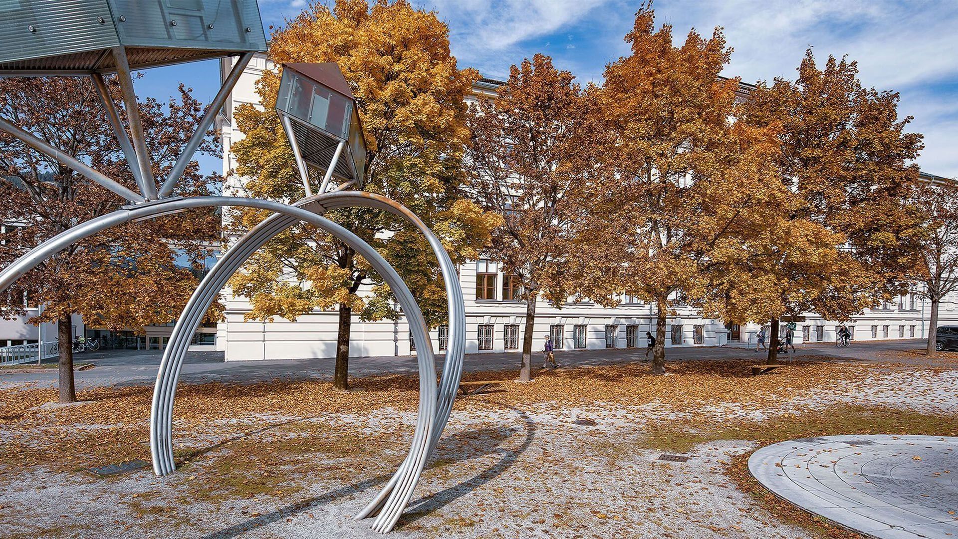 Moderne Skulpturen mit runden Metallbögen in einer Parkanlage, umgeben von Bäumen mit Herbstlaub und einem klaren blauen Himmel.
