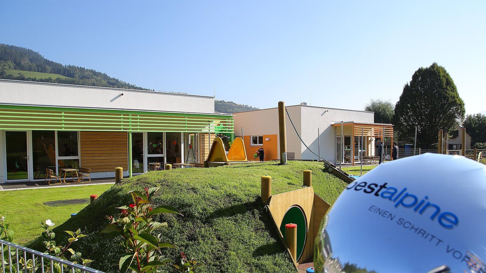 Kindertagesstätte mit Garten, in diesem befinden sich eine Rutsche, ein Spieltunnel sowie kindergerechte Sitzgarnituren