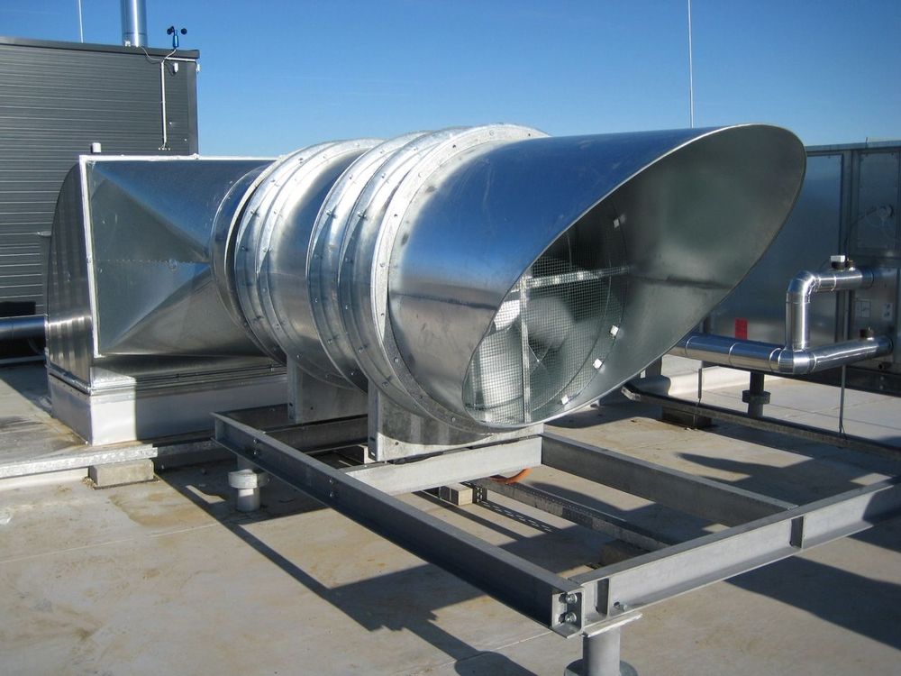 Industrieventilator auf einem Dach, geschützt durch eine Metallabdeckung, umgeben von urbaner Architektur.